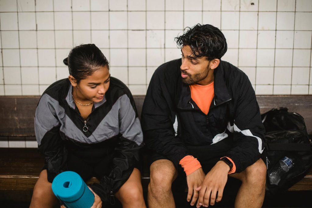 two-young-sport-people-resting-after-fitness-exercises-in-changing-room-5037323 Two people taking a break after fitness training, sitting and chatting indoors.